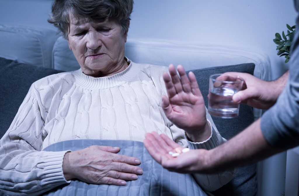 A senior sitting on a couch with a blanket holding their hand up, refusing the medication and glass of water someone got for them to take